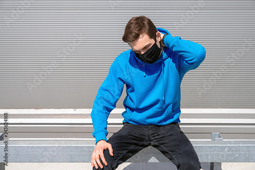 young man in a black mask with an earring, dressed in a blue hoodie, black jeans and black fitness tracker, sits thinking and rubbing the back of his head with his left hand against a gray wall