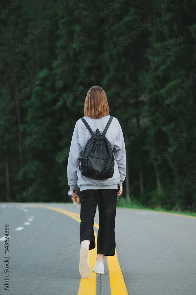 Φωτογραφία Stylish girl in casual clothes and a backpack walking on the asphalted forest road with yellow markings, vertical photo