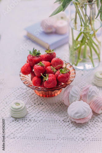 Red ripe strawberries in a glass dish surrounded by green and pink marshmallows, a cup of water and peonies in a glass vase on a white table photo