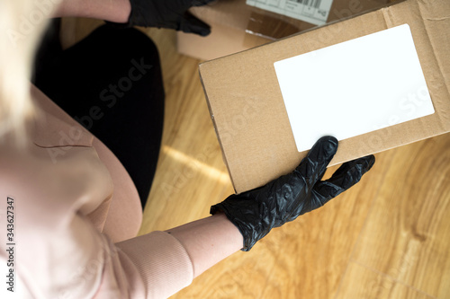 Young blonde girl in protective gloves holding cardboard boxes. Epidemic. Coronavirus. Covid-19. Quarantine. Shipping parcel during coronavirus time. Opening a package during a pandemic in home. © D.