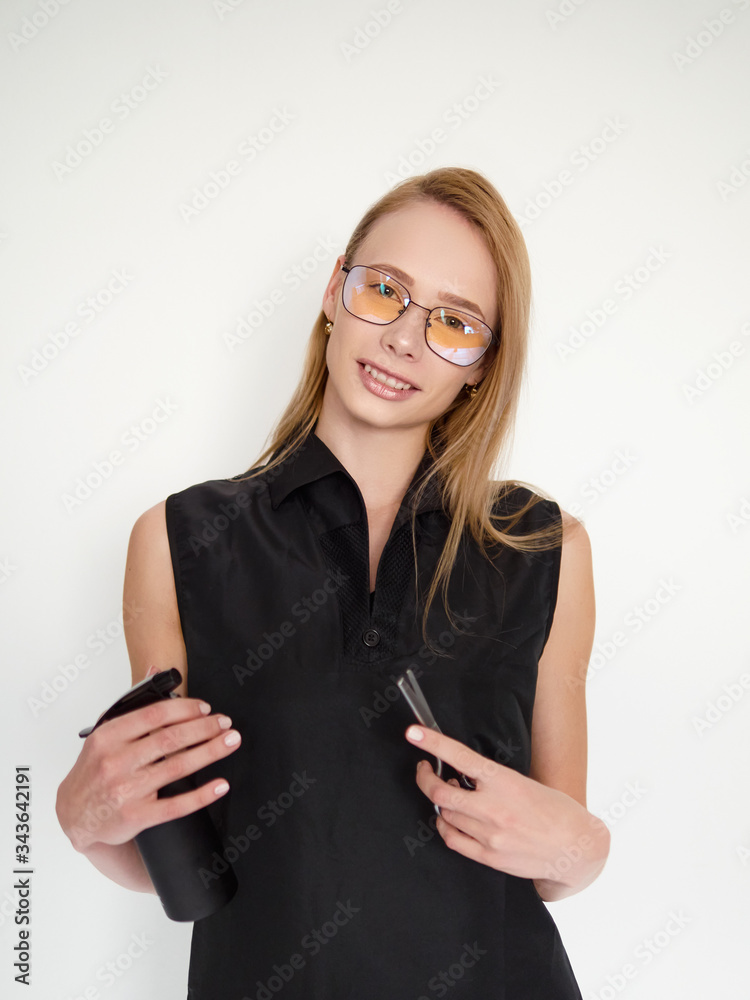 Confident blonde female hairdresser in glasses and black apron standing against white wall in trendy barbershop salon holding professional hair styling tools scissors and spray bottle