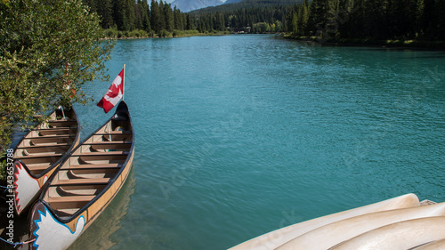 Canoe boat docked by the shore. Turquoise water from the Bow River and Canada Flag background