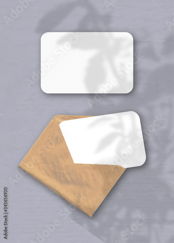 An envelope with two sheets of textured white paper on the gray background of the table. Mockup overlay with the plant shadows. Natural light casts shadows from an exotic plant