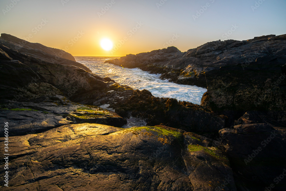 An amazing view of the sunset over the water in the Chilean coast. An idyllic beach scenery with the sunlight illuminating the rocks with orange tones and the sea in the background under a moody sky
