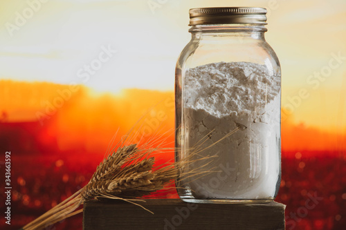 White flour in a glass jar and wheat sheaf with warm sunset farm background. photo