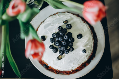 Blueberry cake with white frosting on a white plate next to a vase with 3 fresh pink tulips
