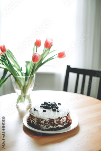 Blueberry cake with white frosting on a white plate next to a clear vase with fresh pink tulips on a wood table with a black simple chair and white curtains in the background