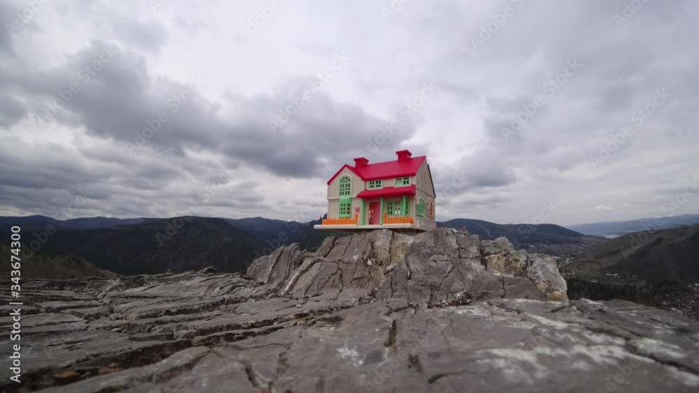 Toy house on stones in a mountain valley. Timelapse. Moving clouds. In background is a village, a green forest. Concept of solitude, relaxation, tourism. Toy house on stones in a mountain valley. Timelapse. Moving clouds. In background is a village, a green forest. Concept of solitude, relaxation, tourism.