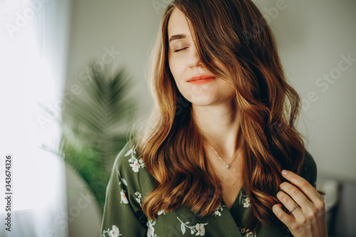 Beautiful young smiling woman with closed eyes in a green patterned dress in a living room with a green plant behind her