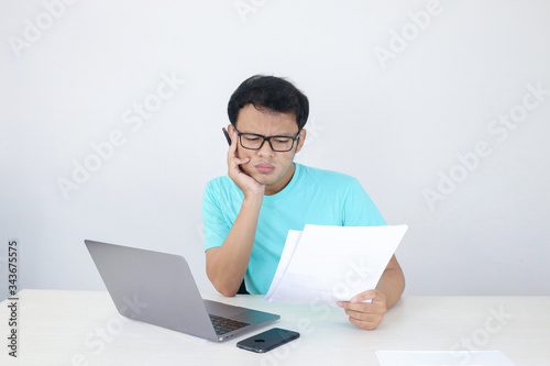 Wow face of Young Asian man shocked what he see at the document and laptop when working isolated grey background. Indonesian man wearing blue shirt.