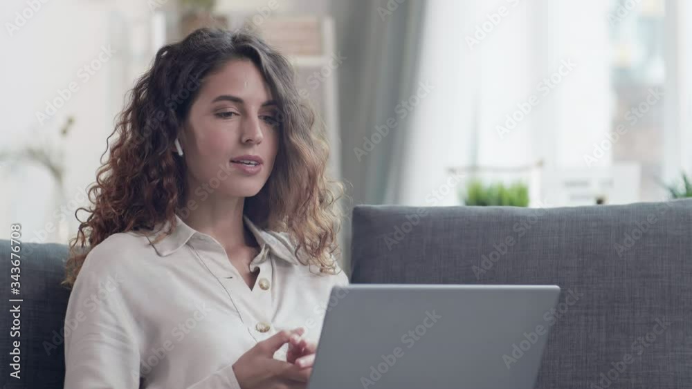 Medium shot of Caucasian curly-haired woman sitting on sofa in living room with laptop on her knees, discussing business strategy with colleague then looking at camera and smiling Medium shot of Caucasian curly-haired woman sitting on sofa in living room with laptop on her knees, discussing business strategy with colleague then looking at camera and smiling