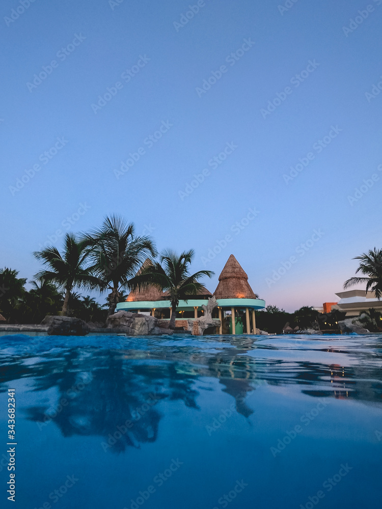 Amazing pool with building with straw roof, palms, and the sunset blue sky, Mexico