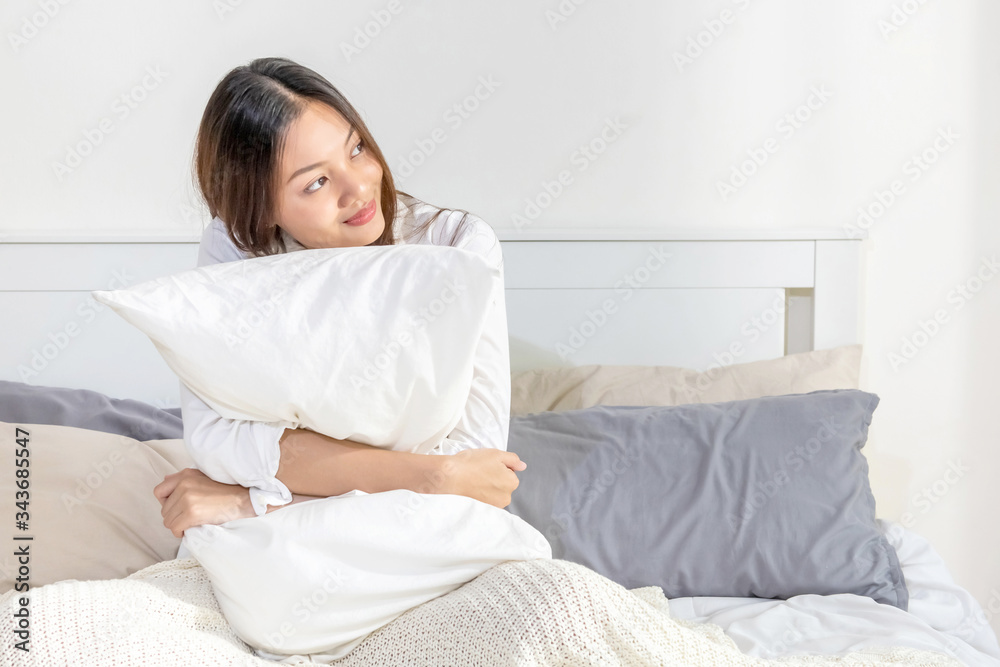 Portrait of smiling relax young beautiful asian woman sitting on the bed with hugging white pillow in bedroom. Happy pretty girl have a good sleep at night and wake up in the morning with freshness. Portrait of smiling relax young beautiful asian woman sitting on the bed with hugging white pillow in bedroom. Happy pretty girl have a good sleep at night and wake up in the morning with freshness.