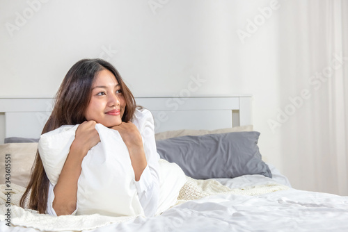 Portrait of smiling relax young beautiful asian woman lying on the bed with hugging white pillow in bedroom. Happy pretty girl have a good sleep at night and wake up in the morning with freshness.