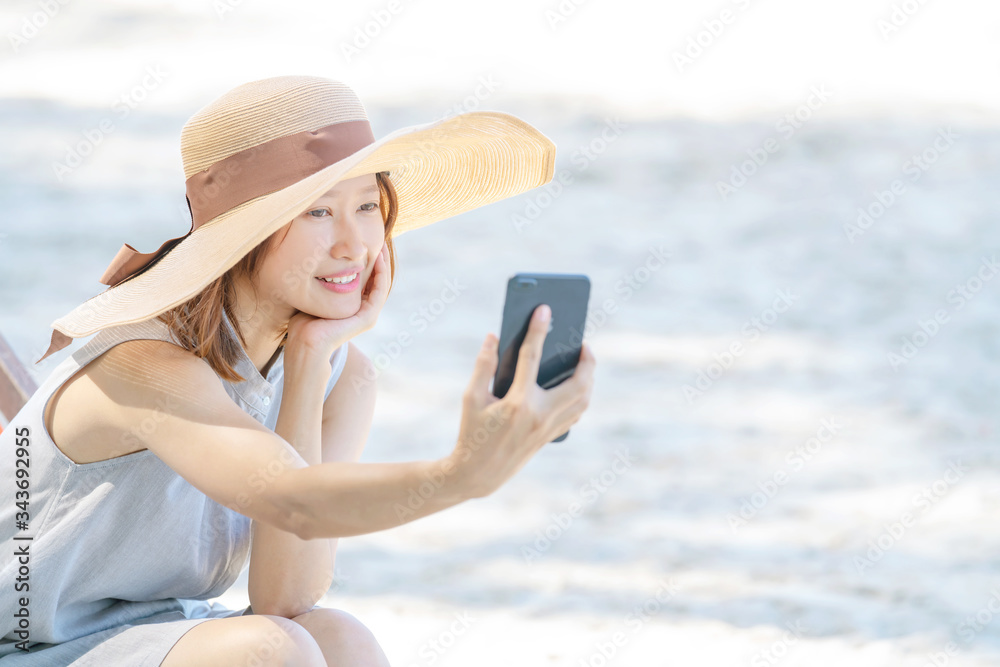 Smiling young beautiful Asian woman girl tourist relax sitting on beach chair at tropical island beach using smartphone selfie or video call or online shopping in sunny summer holiday vacation trip. 