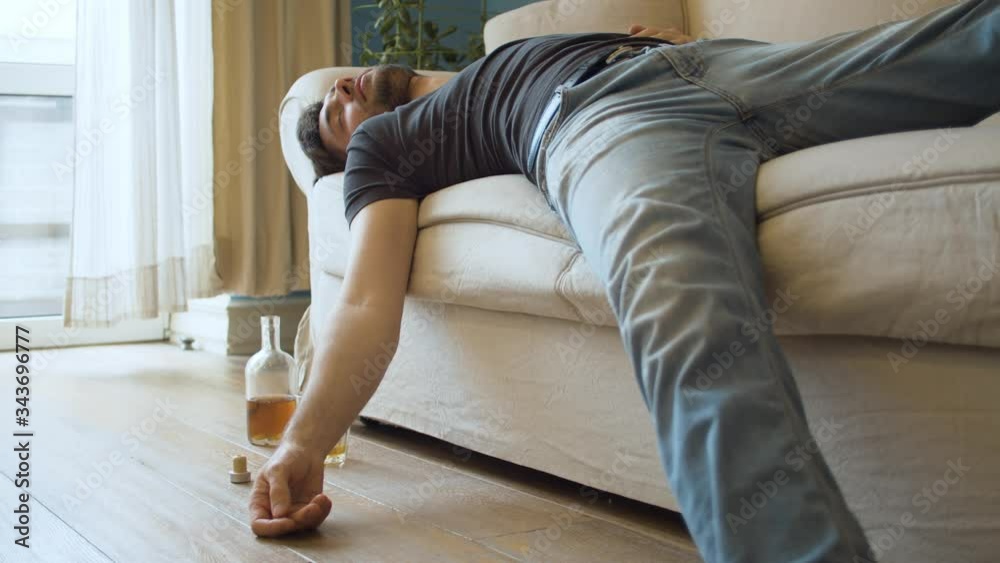 Drunk man sleeping on the couch dressed. Nearby is an unfinished bottle of whiskey and a glass. Young debauchee in his apartment after party Drunk man sleeping on the couch dressed. Nearby is an unfinished bottle of whiskey and a glass. Young debauchee in his apartment after party
