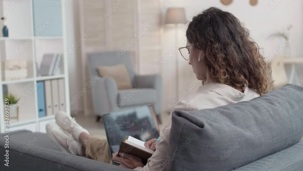 Rear view of Caucasian curly-haired woman with wireless earphones in her ears sitting on couch at laptop, having online lesson and making notes in notebook Rear view of Caucasian curly-haired woman with wireless earphones in her ears sitting on couch at laptop, having online lesson and making notes in notebook