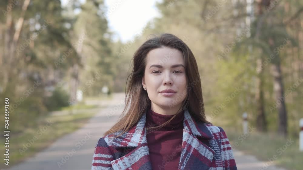 Portrait of confident brunette girl walking to camera along empty small road. Positive brown-eyed Caucasian woman resting next to the forest on sunny day. Leisure, happiness, lifestyle. Portrait of confident brunette girl walking to camera along empty small road. Positive brown-eyed Caucasian woman resting next to the forest on sunny day. Leisure, happiness, lifestyle.