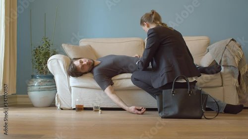 Drunk man sleeping on the couch dressed. His wife in a business suit enters the room. She pulls out his car keys and wallet from his pockets. Young debauchee in his apartment after party
