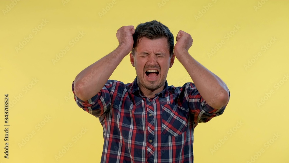 Young man in checkered shirt putting fists on head and screaming in pain. He has a terrible headache. Young man in checkered shirt putting fists on head and screaming in pain. He has a terrible headache.