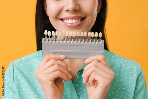 Beautiful young woman with teeth color samples on color background, closeup