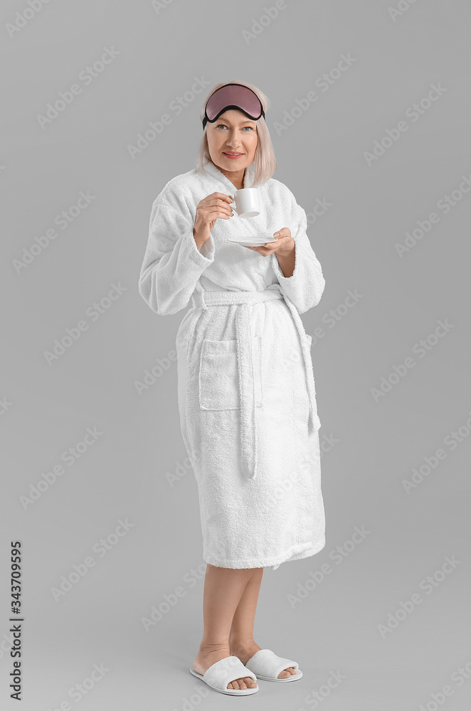 Mature woman in bathrobe, with sleep mask and cup of coffee on grey background
