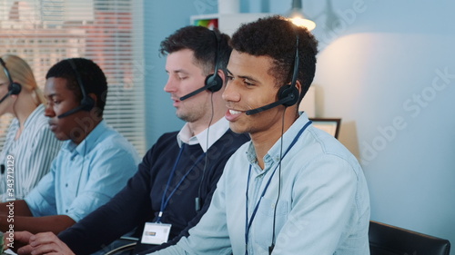 Medium shot of black call center agent feeling exhausted and broken-down after telephone call with the client. He working with mixed-race team in modern office.