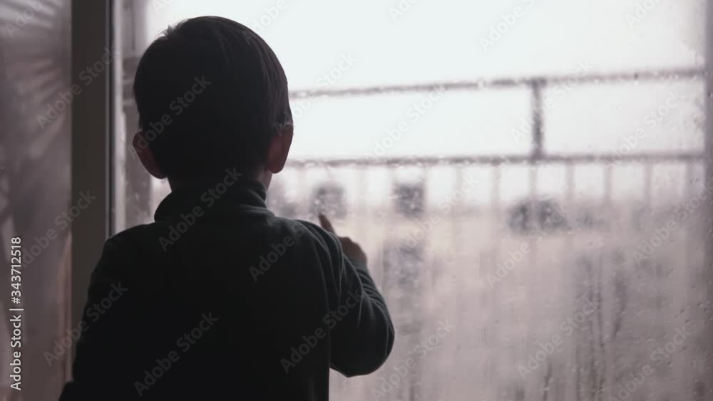 Cute little kid looking and pointing at the street through a glass door covered in condensation and rain drops missing going out during coronavirus lookdown stay home campaign