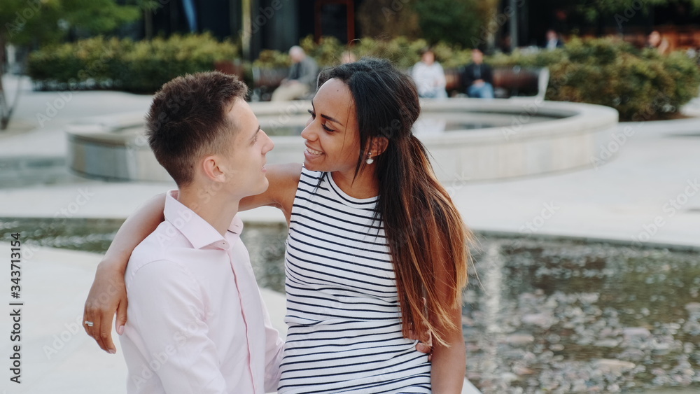Close-up of cheerful multiracial couple having date in a beautiful park. They sitting on the bench, hugging, talking and looking at each other.