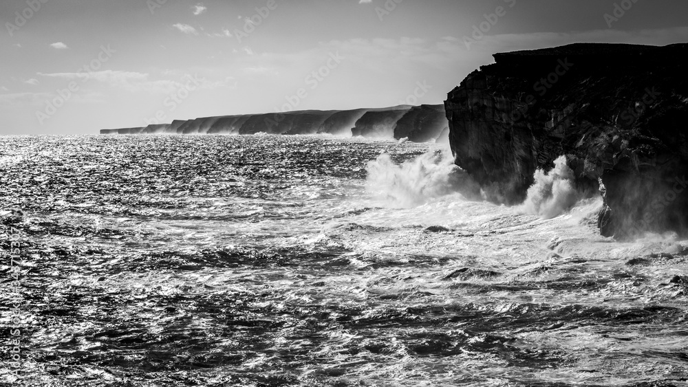 Black and white photo of waves breaking against tall coastal cliffs along the coast line