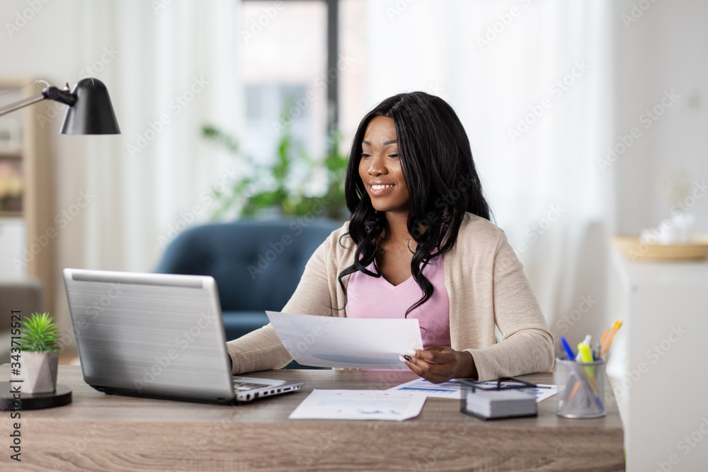 remote job, technology and people concept - happy smiling african american young woman with laptop computer and papers working at home office - obrazy, fototapety, plakaty 