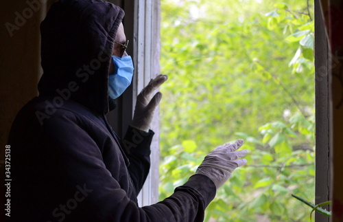 lonely sad man on self-isolation in a dark hoodie, sunglasses, blue surgical antiviral mask and styry gloves touching the glass and looking out the window at the green tree