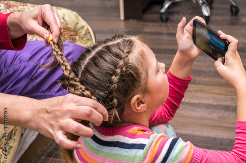 A little girl plays on her phone while her mother does her hair. A woman braids a little girl's hair. A woman combs a child playing in a smartphone.