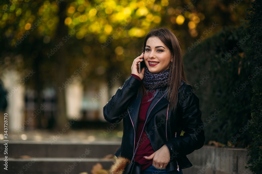 Stylish beautiful lady in a black leather jacket with a black bag and burgundy blouse. Attractive young woman speak by the phone