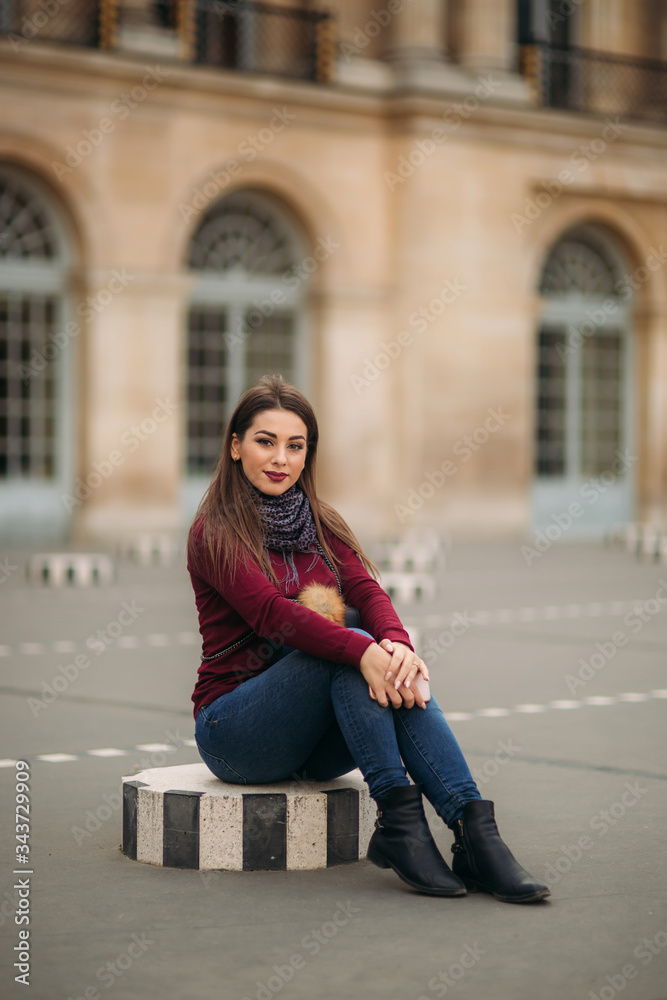 Fototapeta premium Attractive young womanl in burgundy blouse with a gray scarf sits on a striped dais. Happy woman in Paris