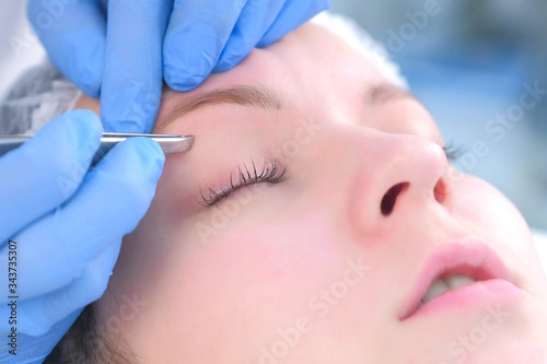 Cosmetologist plucks out hair on eyebrows with tweezers for woman  face closeup. Eyebrows tweezing procedure. Beautician is making brows correction with cosmetic tweezer. Beauty industry.