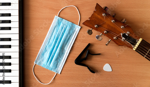 Piano keys, acoustic guitar, capo, pick and blue medical mask lie on a wooden background. Coronavirus epidemic music concept