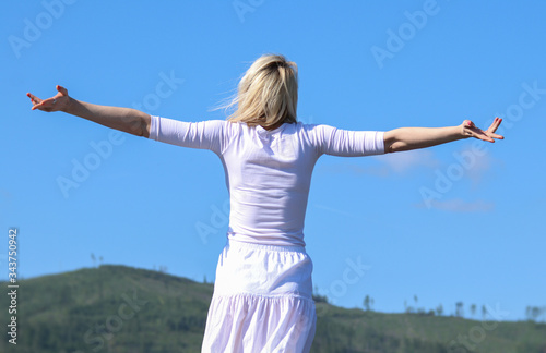 Happy young woman in a white dress spreading her hands in the background of the green hills and the blue sky