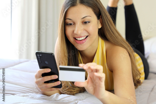 Happy relaxed young woman lying on bed reading her credit card number and enter the number on smart phone doing shopping online comfortable at home. Easy and safe home banking concept.