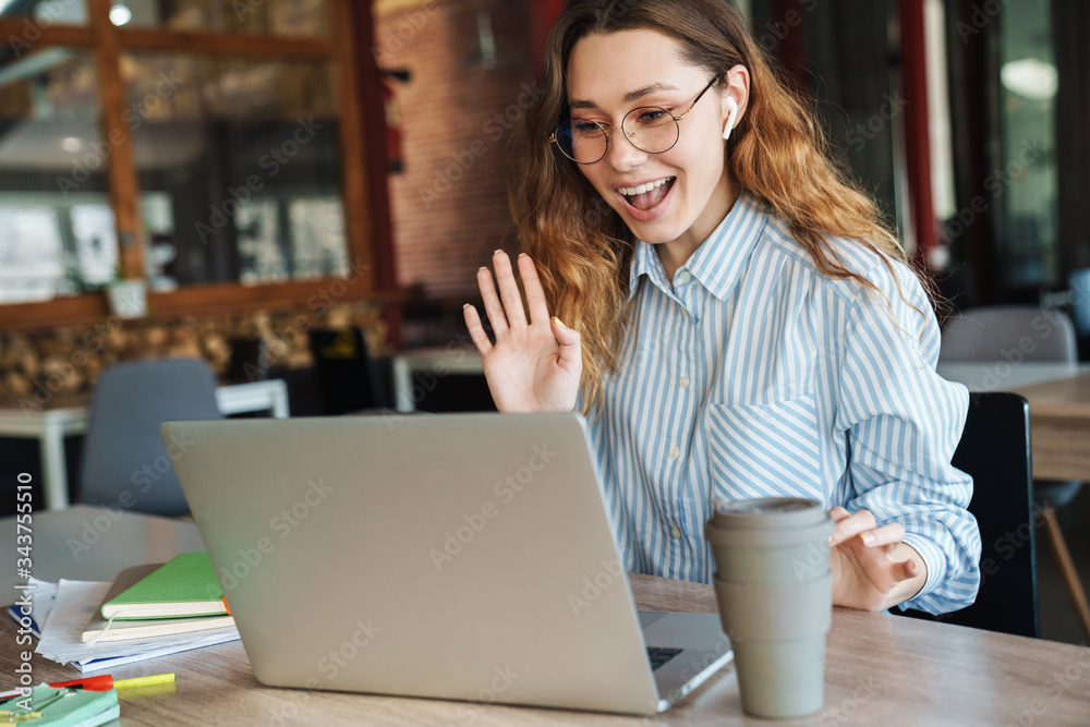 Image of happy woman waving hand and drinking coffee while using laptop Image of happy woman waving hand and drinking coffee while using laptop