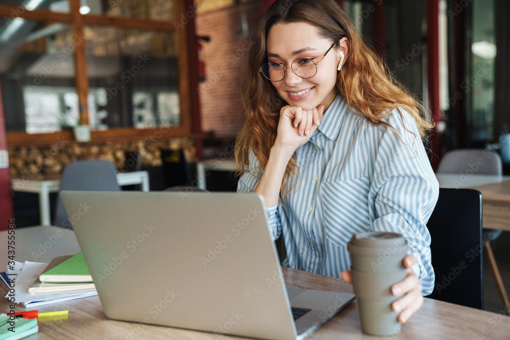 Image of woman drinking coffee while using laptop and wireless earphones