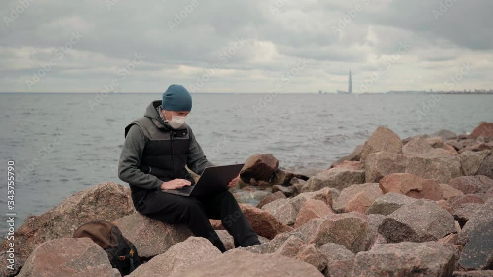 the man in the mask sitting on a rock on the shore of the Bay with the laptop the man in the mask sitting on a rock on the shore of the Bay with the laptop
