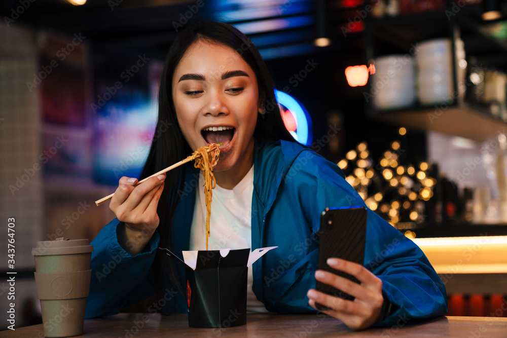 Photo of asian woman eating chinese noodles and using cellphone in cafe