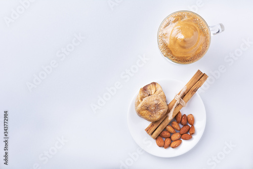 Dalgona Coffee Iced, a Korean fashionable whipped black instant coffee with milk, on a white background. On a white plate with dried figs, almonds, cinnamon sticks. Copy space. Top view.