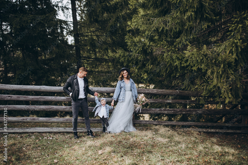 Fototapeta premium Stylish family in the mountains. A guy in a leather jacket, a girl in a wedding dress and a hat with his son stand under a fir tree near a wooden fence and hold a wedding bouquet