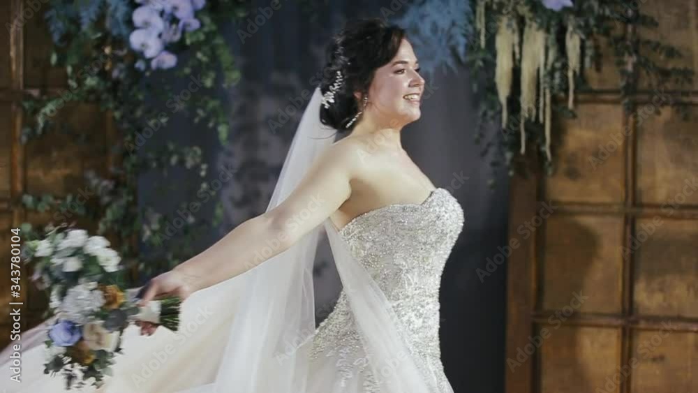Happy charming brunette millennial bride in veil and wedding dress decorated with pearls and rhinestones holds a bouquet of flowers in her hands and swirls against the background of the door and plant