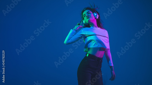 Listening to music. Caucasian young woman's portrait isolated on blue studio background in neon light. Beautiful female model. Concept of human emotions, facial expression, sales, ad, youth culture.