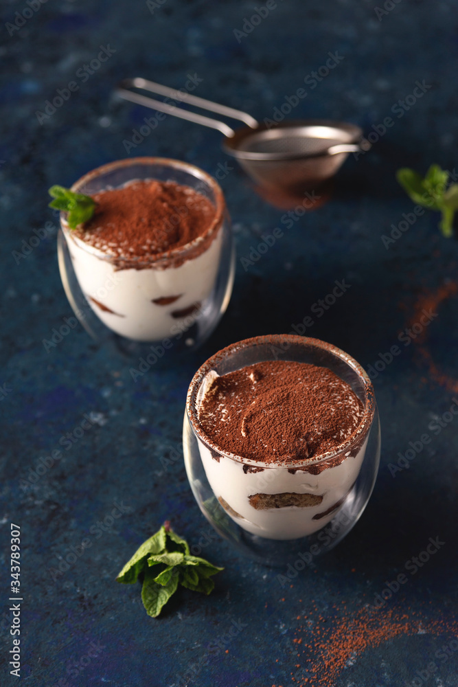 Tiramisu in a transparent glass with a double bottom with cocoa and mint on a blue dark background. Tiramisu in a transparent glass with a double bottom with cocoa and mint on a blue dark background.