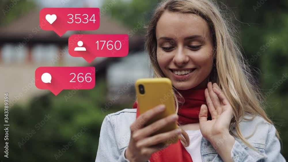 Portrait of young woman wearing in the red scarf using app on smartphone standing near a country house. Animation with user interface with follower, comments, likes counting bubble from smartphone