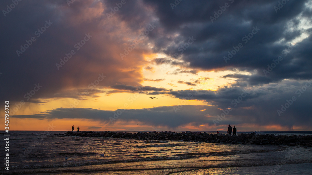Sunset on a sandy beach. Dramatic sky. People on the stone pier.